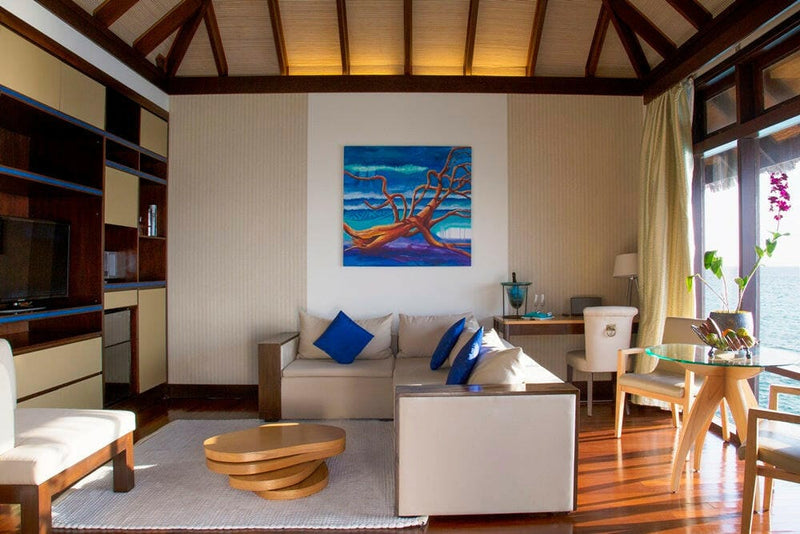 A villa living room at Coco Bodu Hithi, a painted tree canvas above the sofa and shelving along the wall