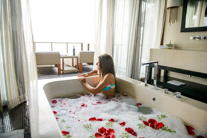 A guest in a bath scattered with red rose petals at Coco Bodu Hithi, the deck loungers through the window