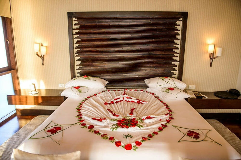A bed laid with red rose petals and folded towels at Coco Bodu Hithi, lit by two wall sconces