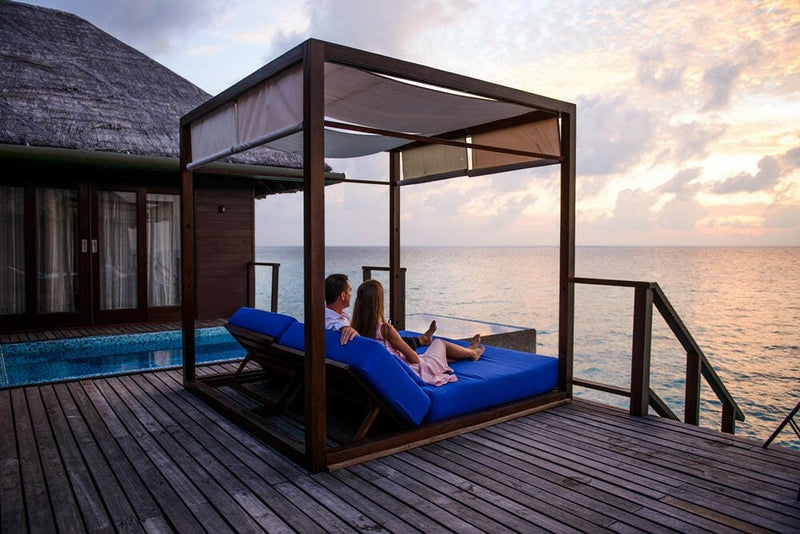 A couple on the canopied deck daybed at Coco Bodu Hithi at dusk, the villa pool beside them