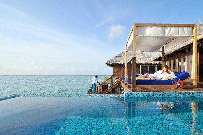 A guest resting on the deck daybed at Coco Bodu Hithi while a host in blue waits at the villa steps