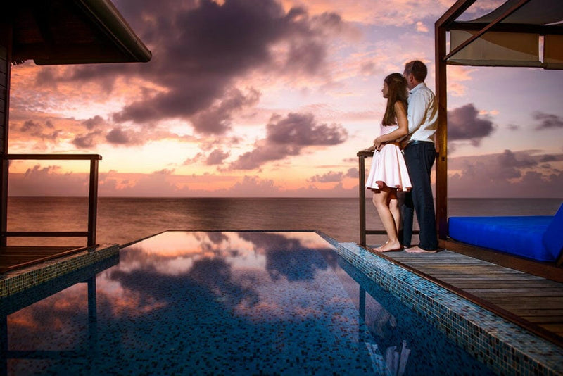 A couple on the villa deck at Coco Bodu Hithi at sunset, pink cloud reflected across the infinity pool