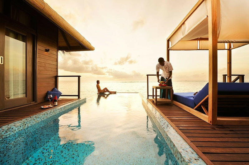 A table being laid on a villa deck at Coco Bodu Hithi at sunset, a guest sitting at the pool edge