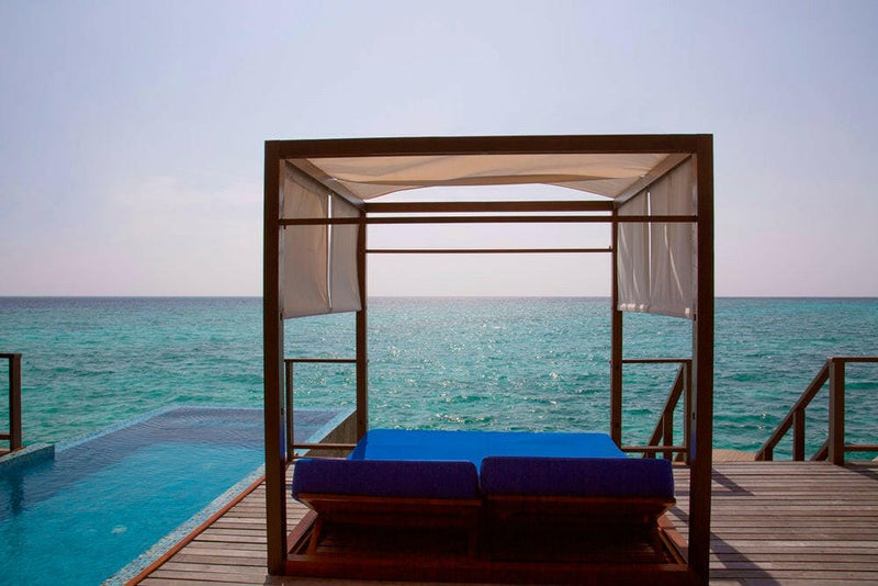 A canopied daybed with a blue mattress at the end of a villa deck at Coco Bodu Hithi, lagoon all round