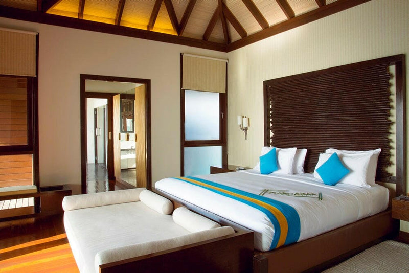 A villa bedroom at Coco Bodu Hithi, turquoise cushions on white linen against a dark slatted headboard