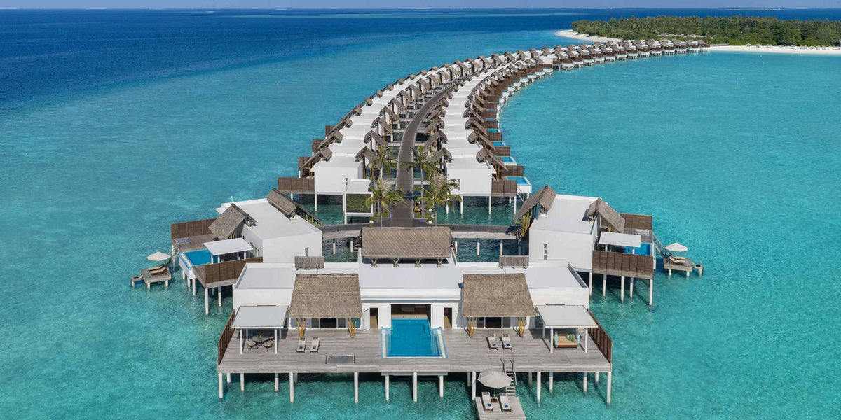 Emerald Maldives Resort & Spa from the air: a long curve of white overwater villas running back to a palm-covered island across pale turquoise shallows.