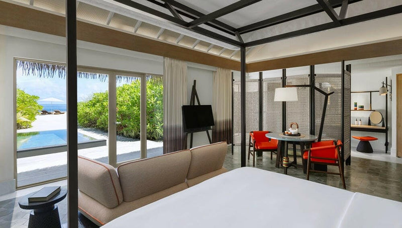 A villa at InterContinental Maamunagau, bed, sofa and red chairs with the pool through glass doors.