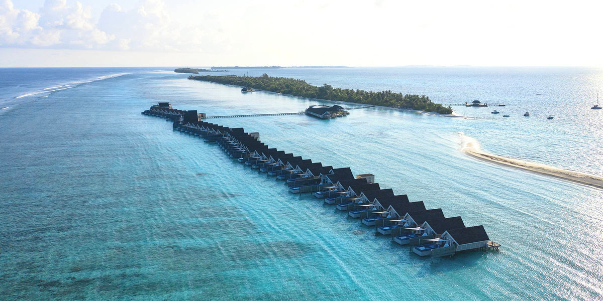 LUX* South Ari Atoll from the air: a long line of overwater villas reaching out from the wooded island across a broad turquoise reef flat, a sandbank alongside.