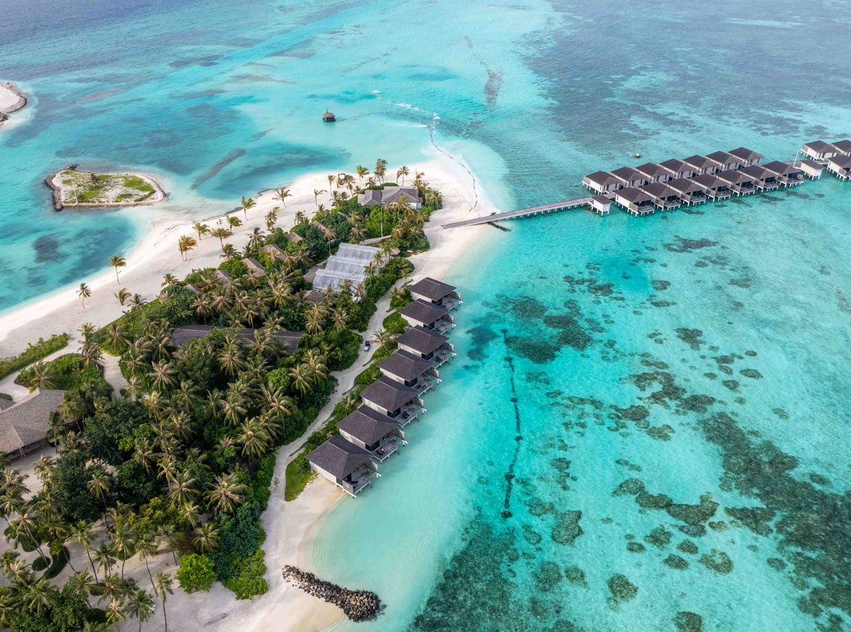 Le Méridien Maldives Resort & Spa from the air, dark-roofed villas among the palms with a jetty across the lagoon