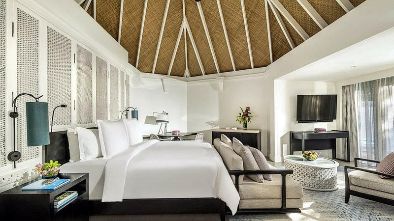 A villa bedroom at Four Seasons Kuda Huraa, a white bed and two armchairs under a high thatched ceiling