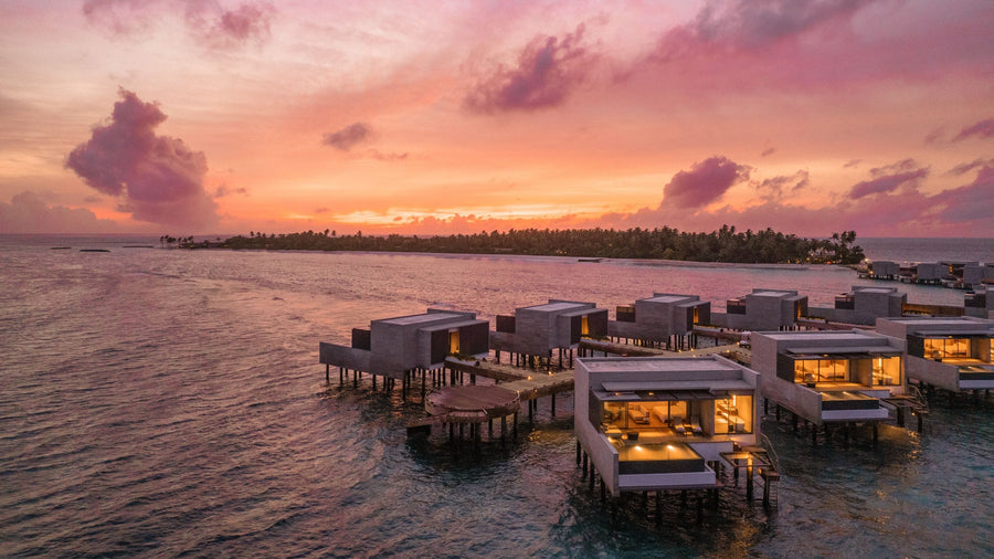 Alila Kothaifaru Maldives, Raa Atoll: overwater villas on stilts lit up at sunset, the island's treeline behind