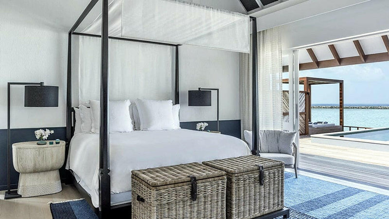 A water villa bedroom at Four Seasons Landaa Giraavaru, a four poster bed and woven trunks facing the deck