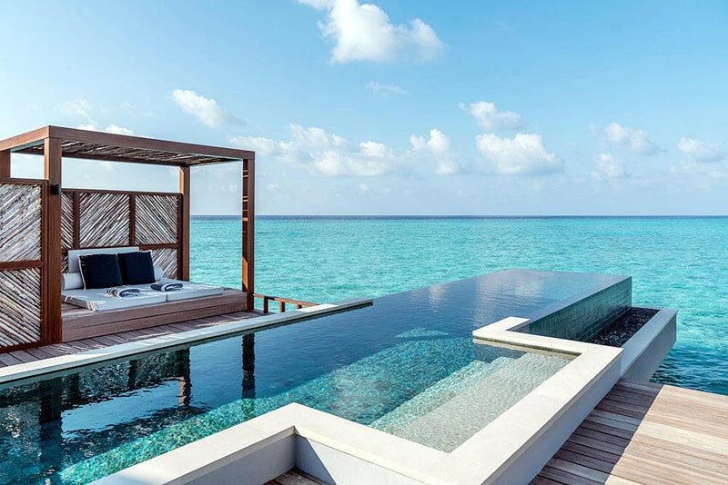 An overwater villa deck at Four Seasons Landaa Giraavaru, an infinity pool and a shaded daybed above the lagoon