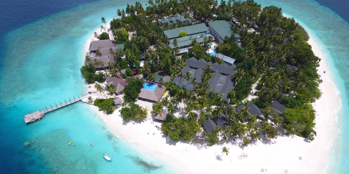 Malahini Kuda Bandos from the air: a small round island densely covered in palms and ringed by white beach, with a jetty on one side and turquoise shallows giving way to deep blue.