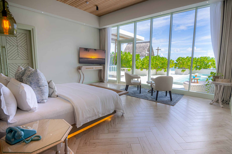 A Beach Pool Suite bedroom at Malahini Kuda Bandos with a herringbone floor and a glass wall onto the terrace and pool.