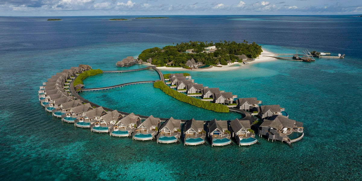 Milaidhoo Maldives from the air: a horseshoe of thatched overwater villas, each with its own plunge pool, curving out from the wooded island across a turquoise lagoon.