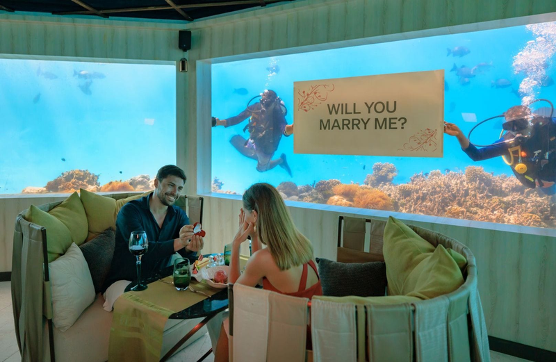 Divers hold up a proposal sign outside the glass of M6m at OZEN LIFE Maadhoo while a couple dines