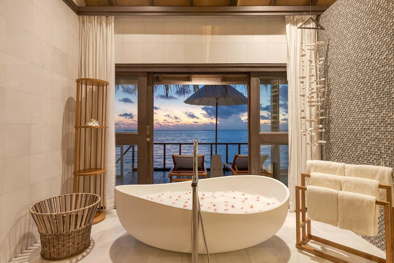 The bathtub of a Wind Villa at OZEN LIFE Maadhoo scattered with petals, the deck and dusk sky through the doors