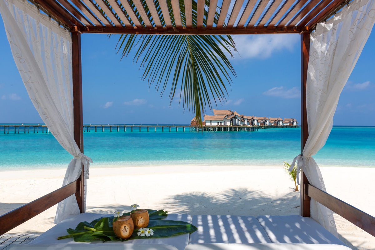 The beach at OZEN Reserve Bolifushi framed by villa curtains, the jetty and overwater villas beyond