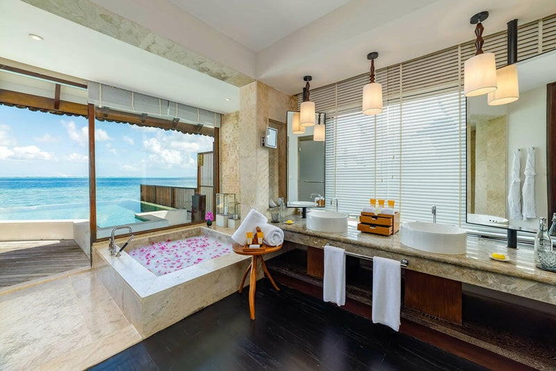 A villa bathroom at OZEN RESERVE Bolifushi, a petal-strewn tub beside twin basins under three pendant lights