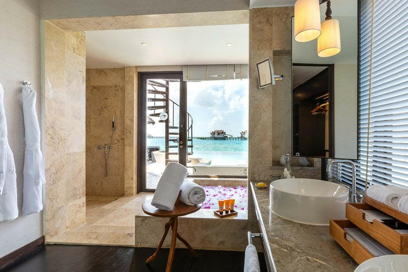 A villa bathroom at OZEN RESERVE Bolifushi, a robe by the shower and a petal-strewn tub facing the sea