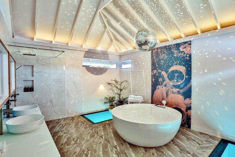 A villa bathroom at The Standard Huruvalhi, an oval tub below a mirror ball beside a flamingo mural.