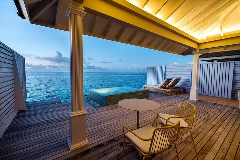 A water villa deck at The Standard Huruvalhi, a round table and tan chairs beside a lit timber roof.