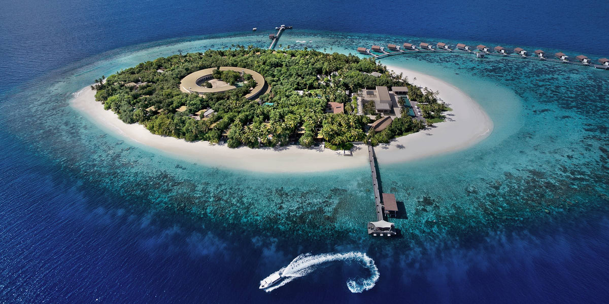 Park Hyatt Maldives Hadahaa from the air: a small round wooded island ringed by white sand and reef, a jetty with a speedboat leaving a wake, and overwater villas along the far shore.