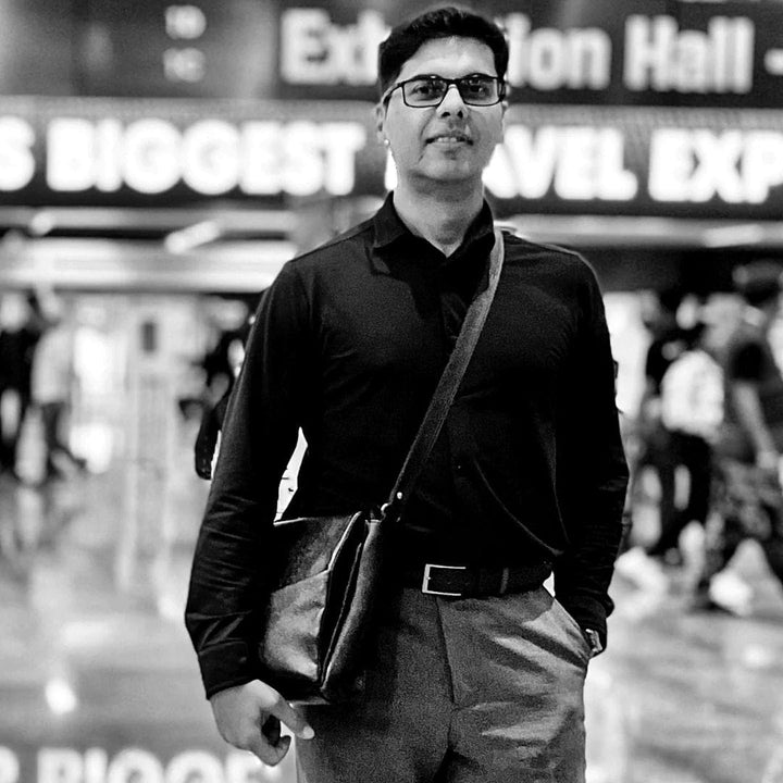 A black and white portrait of Pranjal Sarmah wearing glasses, a dark long-sleeve polo shirt, and trousers, carrying a shoulder bag, at a travel event.