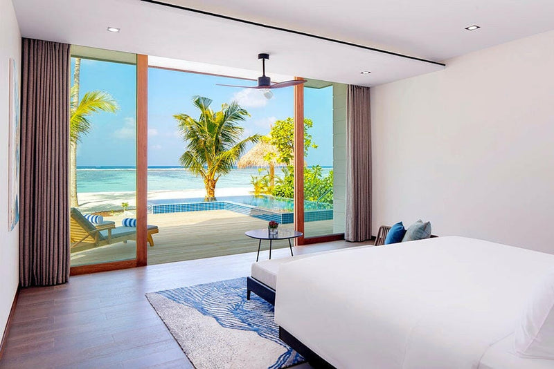 A villa bedroom at Radisson Blu Maldives, timber doors sliding open onto a pool terrace and palms.
