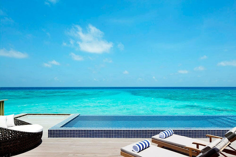 A villa deck at Radisson Blu Maldives, pool edge meeting the sea, wicker daybed and rolled towels.