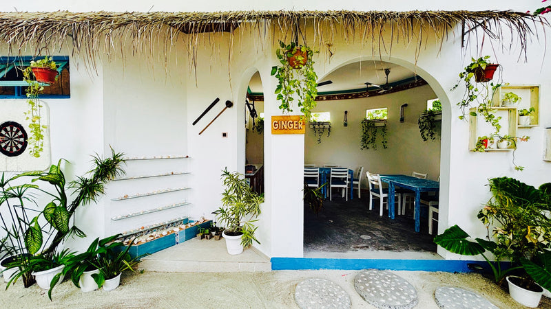Entrance to the Ginger restaurant at Dream Inn Thulusdhoo, a whitewashed building with a thatched roof, potted plants and blue tables inside