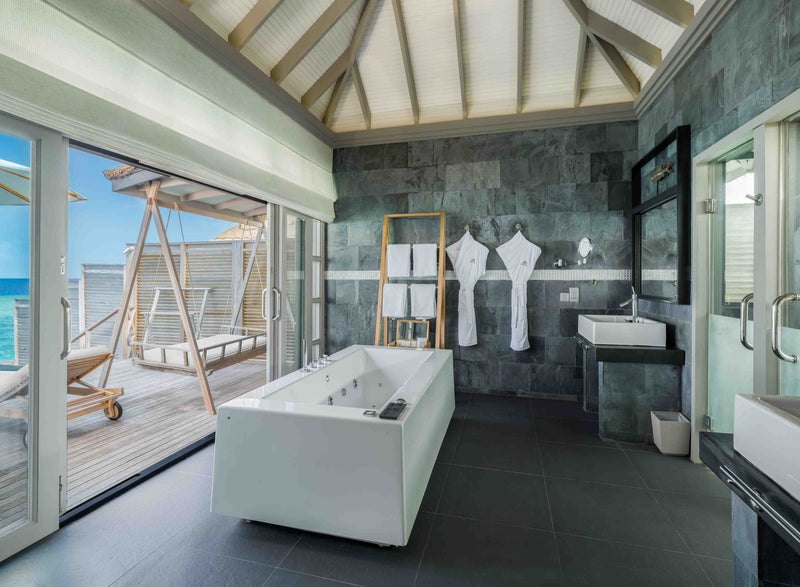 A water villa bathroom at JA Manafaru, a freestanding tub against grey stone with doors open to the deck.