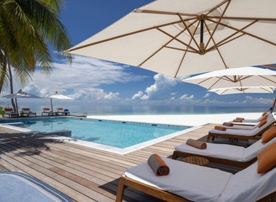 The pool deck at V Villas Maldives at Mirihi, loungers under white parasols beside the pool with the beach and sea beyond.
