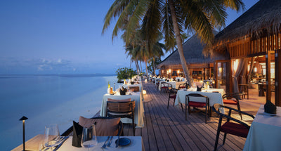 Tables laid along a timber deck beside the beach at V Villas Maldives at Mirihi at dusk, palms overhead and the lagoon alongside.