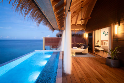 A villa deck at V Villas Maldives at Mirihi at dusk, the pool lit beside a daybed with the bedroom open behind.