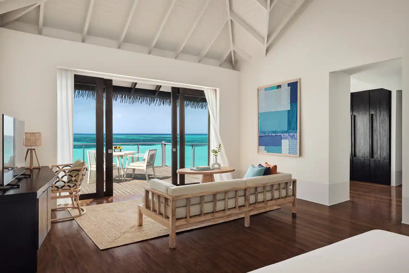 A water villa interior at Noku Maldives, rattan sofa with teal cushions facing doors to the sea.