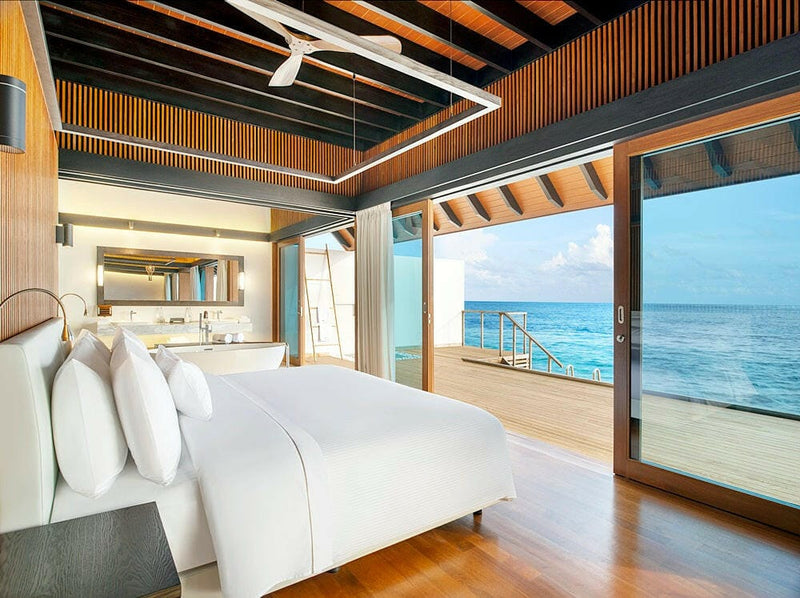 A white bed at The Westin Maldives Miriandhoo, glass doors open to a deck above the reef.