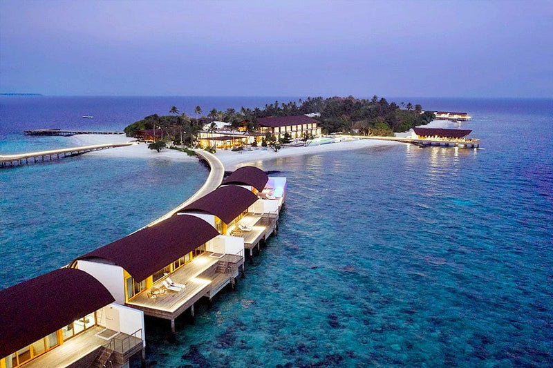 The Westin Maldives Miriandhoo at dusk, lit overwater villas curving away from the island.