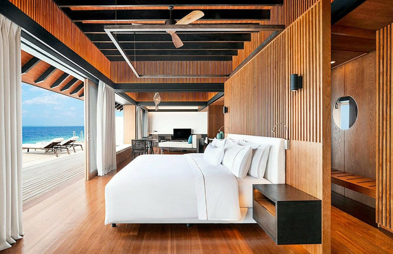A bedroom at The Westin Maldives Miriandhoo, timber walls, oval mirror and a lounger on the deck outside.