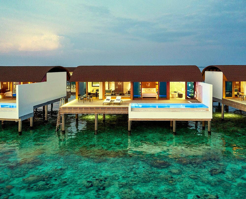 Overwater villas at The Westin Maldives Miriandhoo at dusk, warm light in the rooms and lit pools.