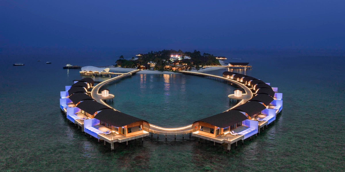 The Westin Maldives Miriandhoo Resort at dusk, seen from the air: a ring of lit overwater villas curving around the lagoon.
