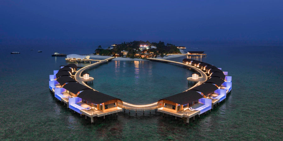 The Westin Maldives Miriandhoo Resort at dusk, seen from the air: a ring of lit overwater villas curving around the lagoon, with the wooded island and its jetty behind.