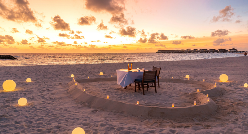 A private dinner table at Avani+ Fares Maldives, inside a heart drawn in the sand with lanterns lit at sunset.