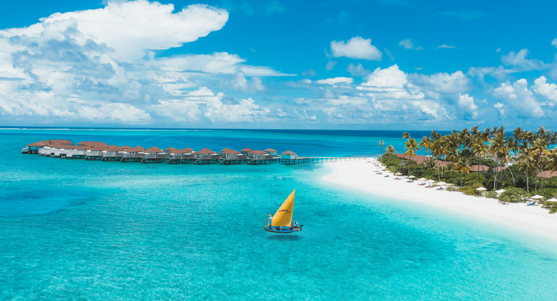 A dhoni with a yellow sail at Avani+ Fares Maldives, passing the overwater villas near a sandbank.