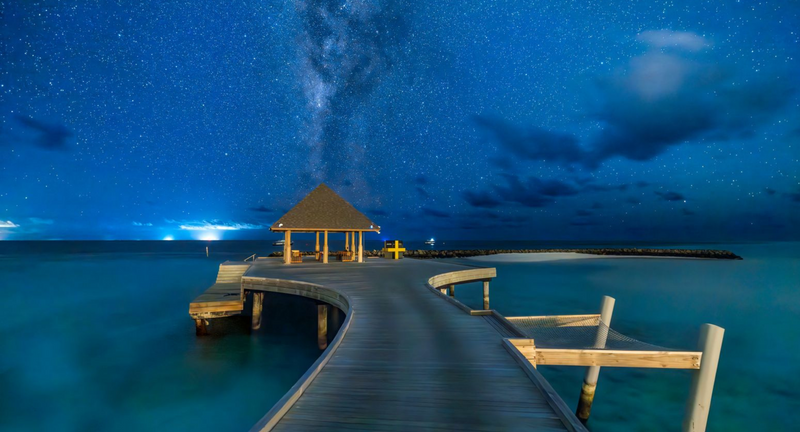 The arrival jetty at Avani+ Fares Maldives, its thatched pavilion under a starry night sky.