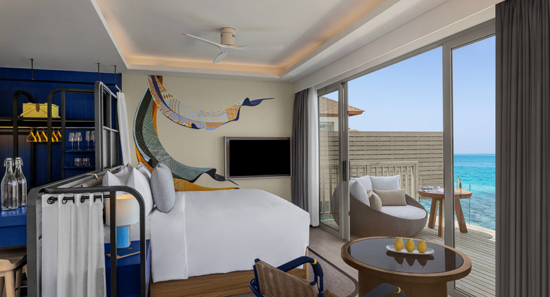 An Overwater Villa bedroom at Avani+ Fares Maldives, in blue and white, with glass doors to a balcony over the sea.