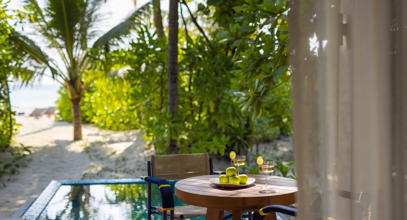 A shaded Beach Pool Villa terrace at Avani+ Fares Maldives, a table set with fruit and the sand and palms beyond.