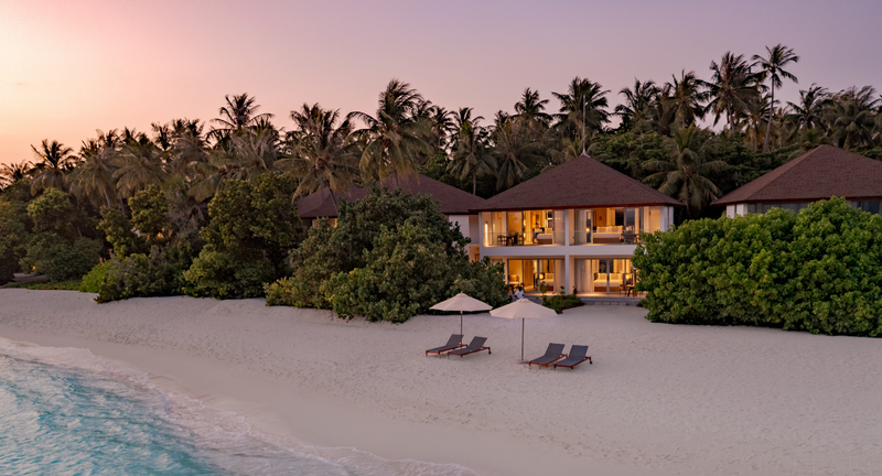 Two-storey beachside buildings at Avani+ Fares Maldives, behind the sand at sunset with loungers in front.