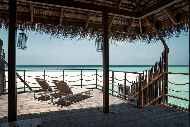 Constance Moofushi, South Ari Atoll: covered deck of a Senior Water Villa with loungers and a view across the lagoon to the beach